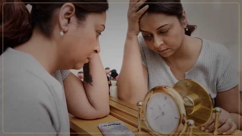 A tired woman rests her head beside a mirror and clock, representing the cumulative biological cost of chronic stress over time